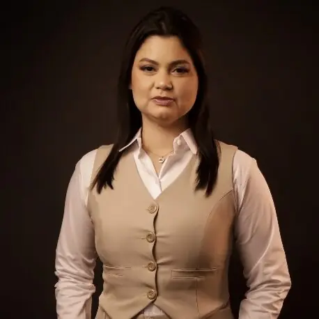 Retrato de Gesiane Gonçalves de colete bege sobre camisa clara, braços ao lado do corpo, em estúdio de fundo marrom-escuro